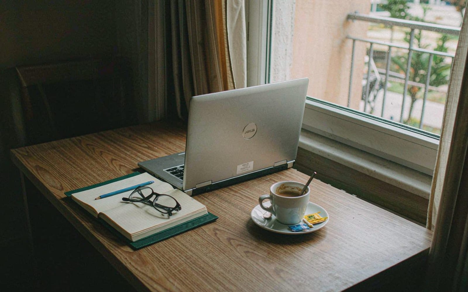Caderno, laptop e xícara sobre mesa junto à janela
